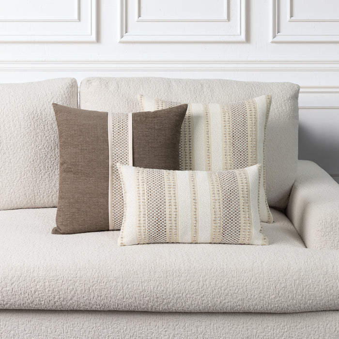 Three decorative throw pillows in neutral tones of brown, beige, and cream, styled on a textured off-white boucle sofa against a white paneled wall.