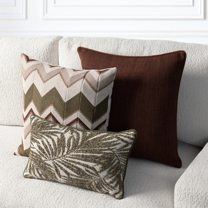 Three decorative throw pillows arranged on a light beige textured sofa: a large dark brown pillow, a square chevron-patterned pillow in beige, brown, and olive green, and a smaller rectangular pillow with a brown and beige leaf pattern.