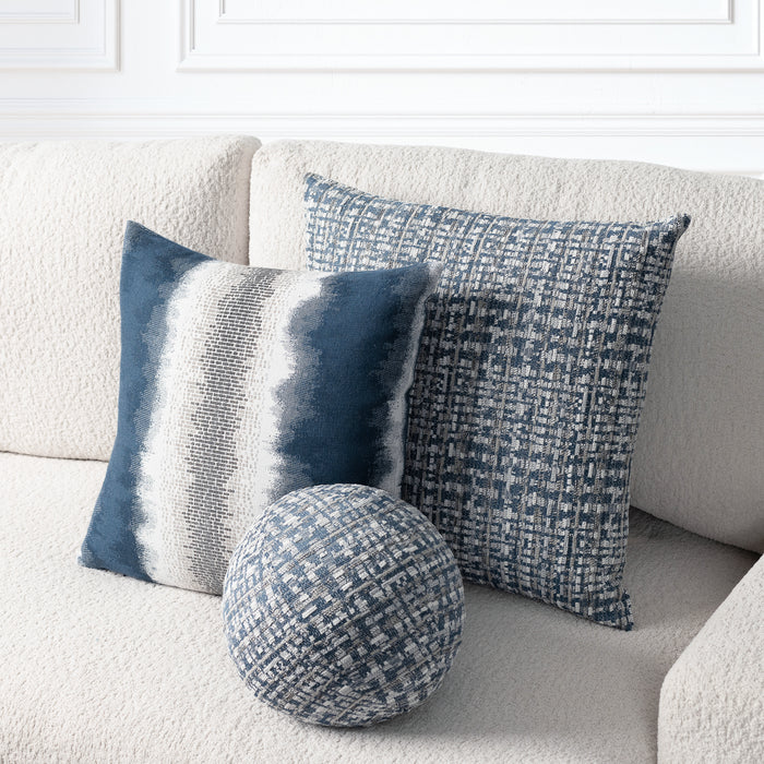 Three decorative throw pillows in blue, navy, and gray tones arranged on a textured cream sofa. The set includes two square pillows with woven patterns and one round matching accent pillow.