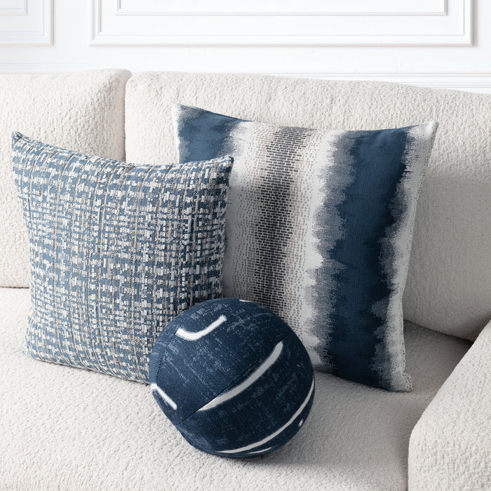 Three decorative throw pillows arranged on a cream textured sofa: a square pillow with a blue-and-white woven pattern, a square pillow with vertical blue, gray, and white brushstroke stripes, and a round navy pillow with white curved stripes. White wall molding is visible in the background.