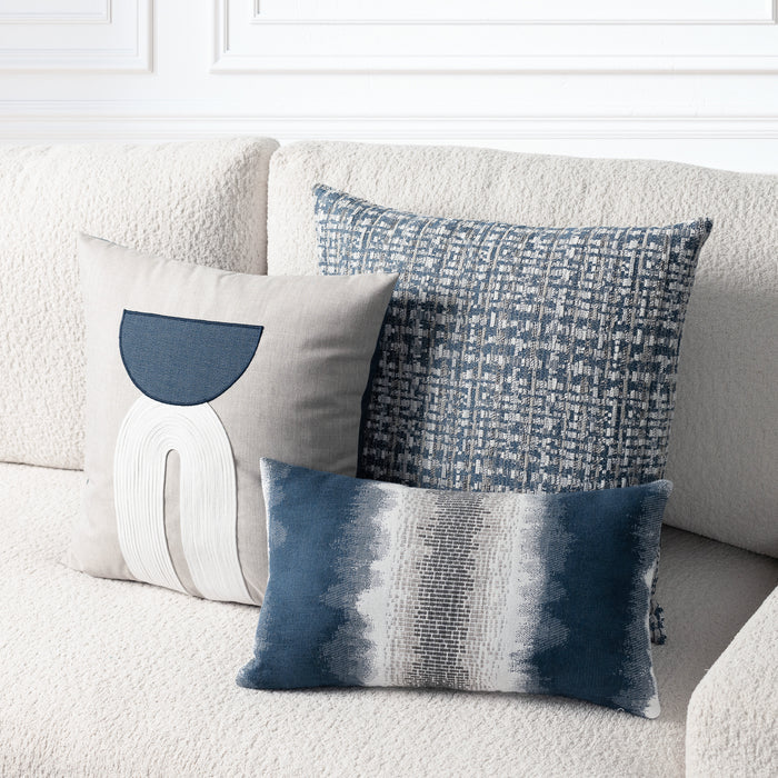 Three decorative throw pillows arranged on a cream boucle sofa against a white paneled wall. The pillows include a light beige pillow with a dark blue semicircle and white curved line motif, a blue-and-white patterned square pillow, and a horizontal rectangular pillow with a blue-to-white gradient texture.