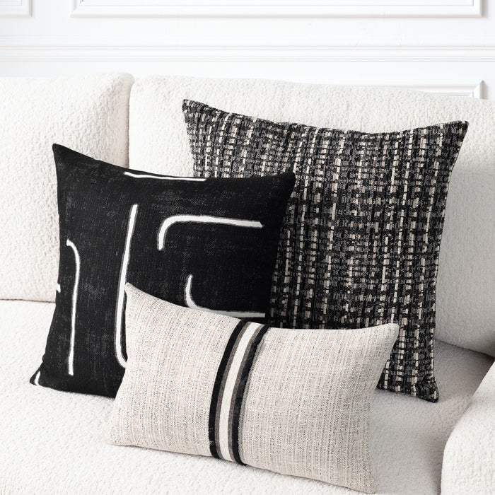 Three decorative throw pillows arranged on a textured off-white sofa. The pillows include a black square with white abstract lines, a black and gray woven square, and a light beige rectangular pillow with two black stripes.