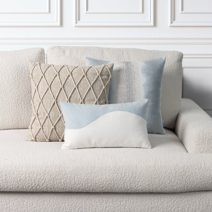 Three decorative throw pillows arranged on a textured cream boucle sofa against a white paneled wall. The pillows include a square beige woven diamond-pattern pillow, a light blue watercolor-style square pillow, a small white pillow with a light blue curved top, and a larger light blue rectangular pillow.