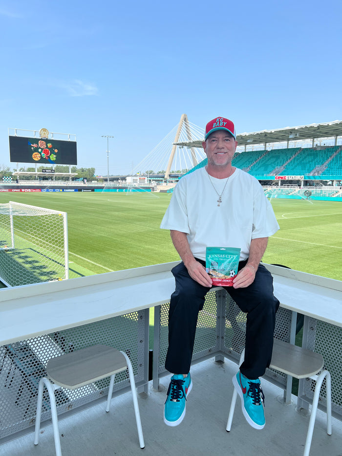 A man sitting on a white stadium bench overlooking a green soccer field, wearing a white t-shirt, black pants, and bright blue sneakers, holding a red and white packaged product.