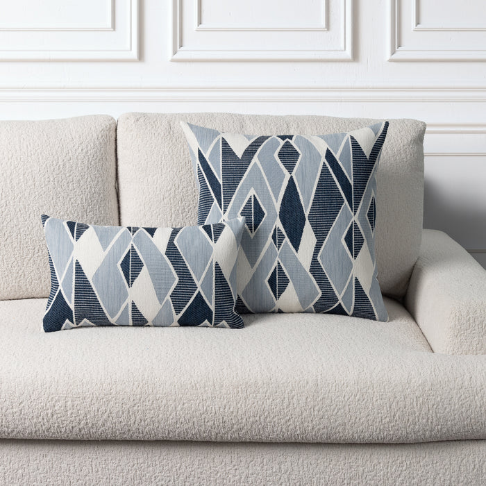 Two decorative throw pillows with a geometric diamond pattern in navy, light blue, and white, styled on a textured cream boucle sofa against a white paneled wall.