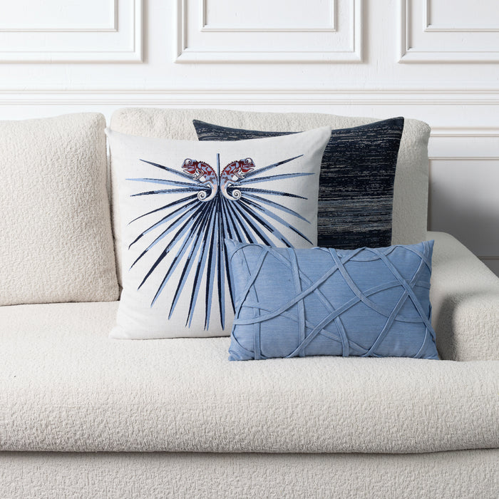 Three decorative throw pillows arranged on a cream bouclé sofa against a white paneled wall. The front pillow is white with a blue fanburst motif and red floral accents, the middle pillow is dark blue-gray with a textured vertical stripe, and the front pillow in front is light blue with a raised lattice pattern.