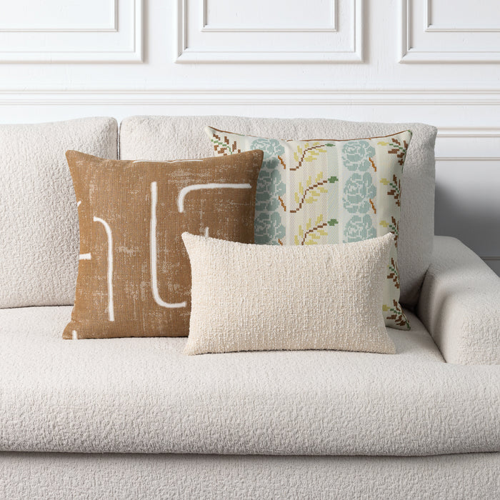 Three decorative throw pillows arranged on a light beige boucle sofa against a white paneled wall. The front pillow is a textured cream rectangle, the center-left pillow is tan linen with a white abstract line design, and the back pillow has a muted floral pattern in sage green, beige, and brown.