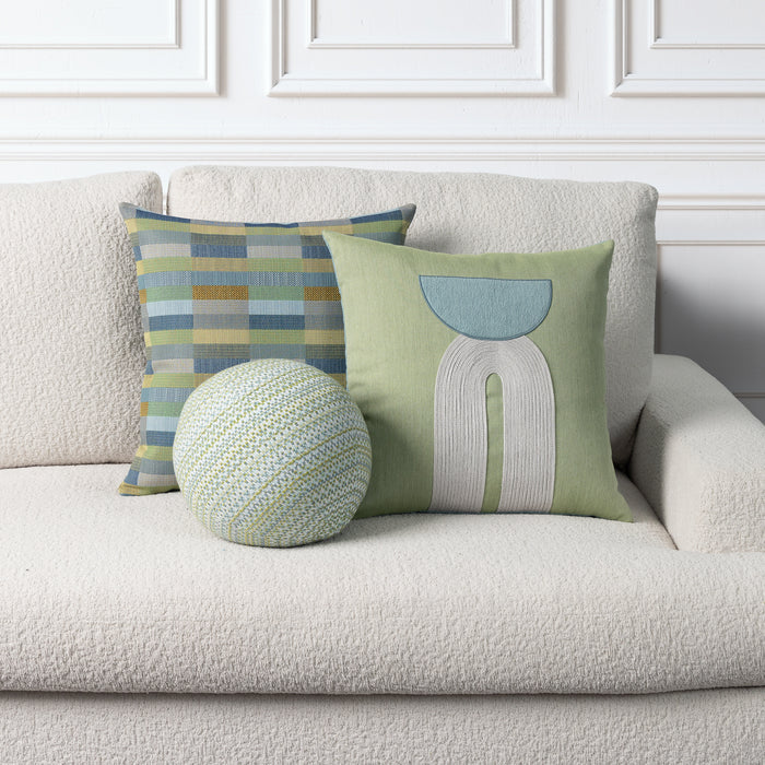Three decorative throw pillows arranged on a textured cream boucle sofa against a white paneled wall. One square pillow has a green base with a light blue and white abstract arch design, one square pillow has a blue, green, and beige striped pattern, and one round pillow is knitted in light green and cream.