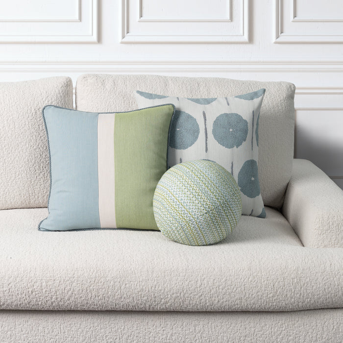 Three decorative throw pillows arranged on a textured cream boucle sofa against a white paneled wall. One square pillow is light blue and sage green with a white stripe; one square pillow has a white background with blue-gray circular motifs; one round pillow is green, blue, and white patterned.