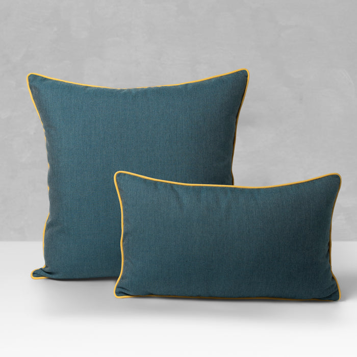 Two teal throw pillows with mustard yellow piping, one square and one rectangular, displayed on a white surface against a textured gray background.