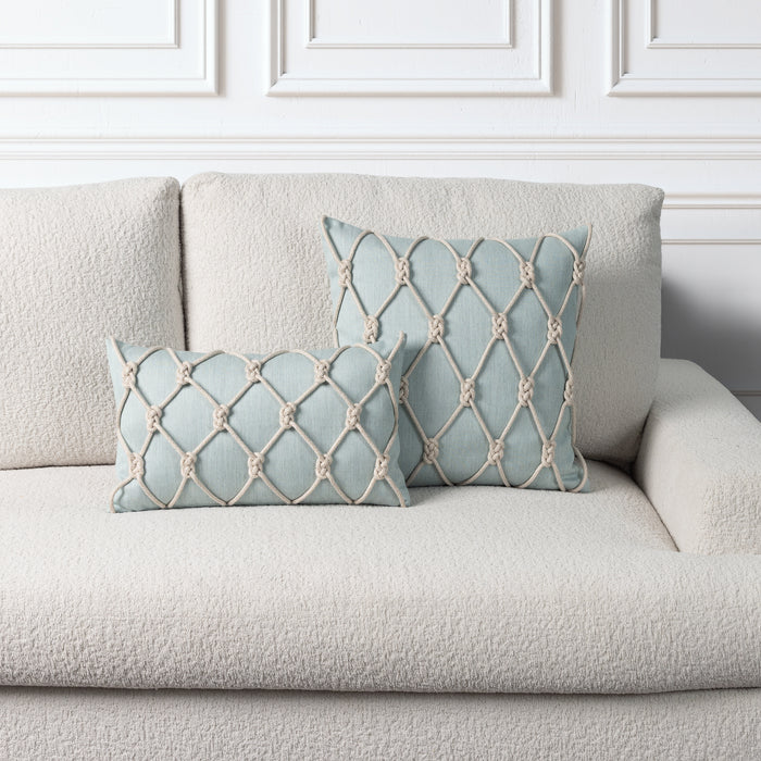 Two light blue decorative throw pillows with cream rope-knotted geometric patterns, displayed on a textured cream bouclé sofa against a white paneled wall.