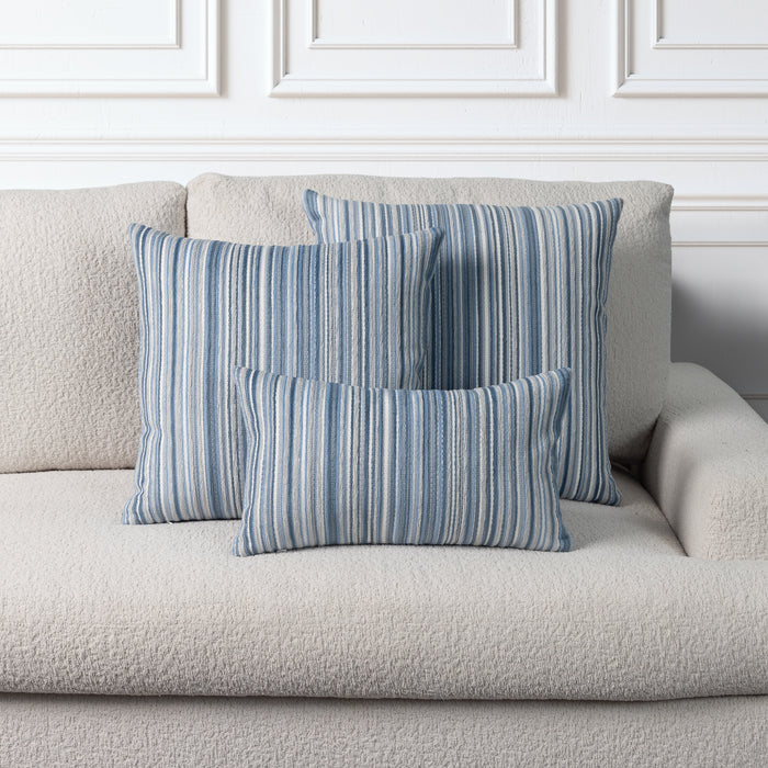 Three striped throw pillows in shades of blue, navy, and white, arranged on a textured light beige sofa against a white paneled wall.
