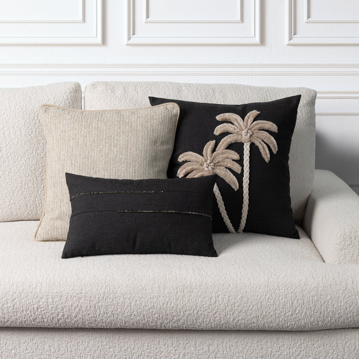 Three decorative throw pillows arranged on a textured cream boucle sofa against a white paneled wall. The set includes a large black pillow with beige rope-style palm tree embroidery, a smaller black pillow with horizontal gold metallic stitching, and a neutral beige woven pillow.