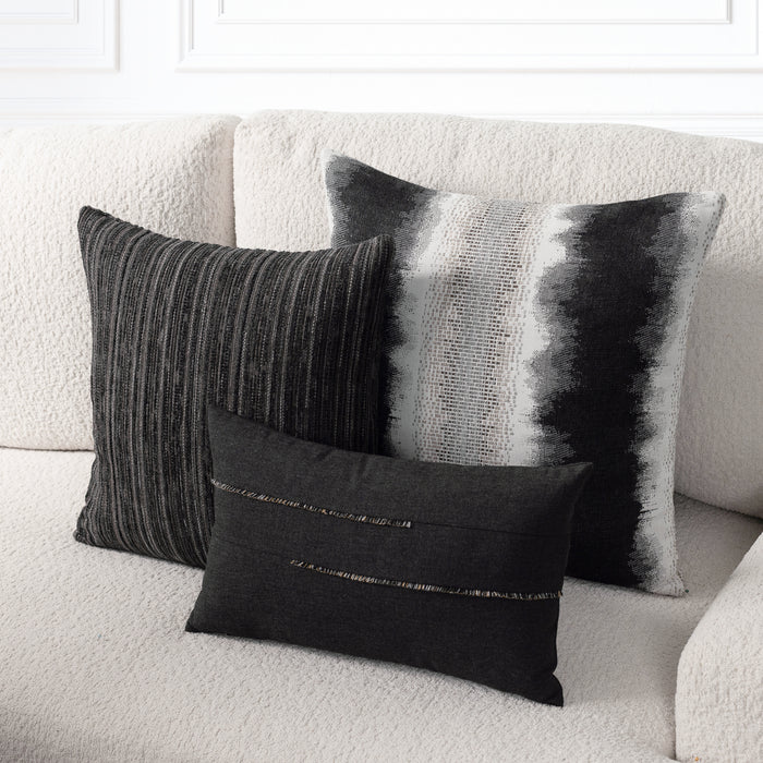 Three decorative throw pillows in black, gray, and white tones arranged on a textured cream sofa. One pillow has vertical gray stripes, one has a black and white brushstroke pattern, and the front pillow is black with a thin metallic horizontal stripe.