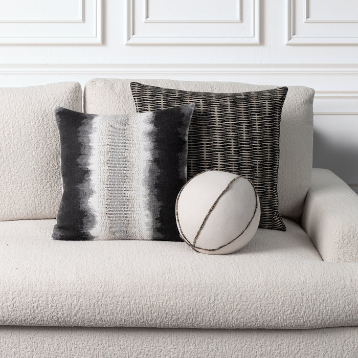 Three decorative throw pillows arranged on a light beige textured sofa against a white paneled wall. One square pillow has vertical black, gray, and white stripes, one square pillow has a black-and-white woven pattern, and one round pillow is off-white with dark stitched lines.