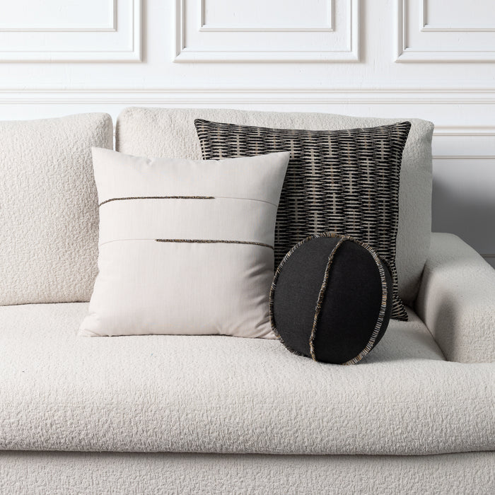 Three decorative throw pillows arranged on a light beige textured sofa against a white paneled wall. The set includes a large cream pillow with two horizontal black stitched lines, a black and gray woven patterned pillow, and a small round black lumbar pillow with a fringed edge.