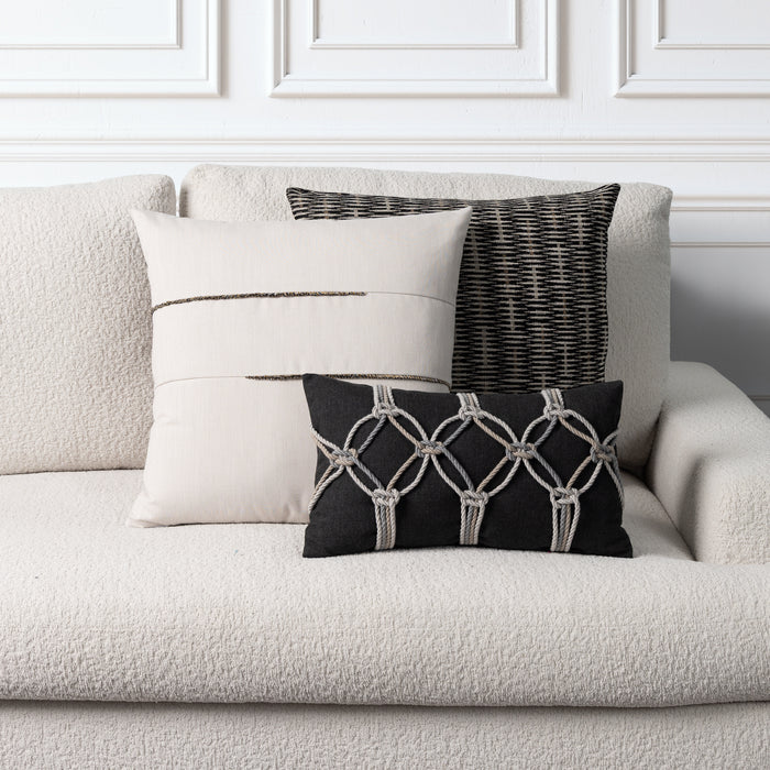 Three decorative throw pillows arranged on a textured light beige sofa against a white paneled wall. The pillows include a large cream pillow with a thin dark stripe, a black-and-white woven vertical pillow, and a black lumbar pillow with a cream geometric knot pattern.
