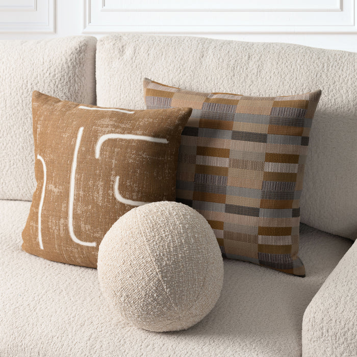 Three decorative throw pillows arranged on a textured cream sofa: a square tan pillow with white abstract line art, a square plaid pillow in tan, brown, and gray, and a round cream textured pillow in front.