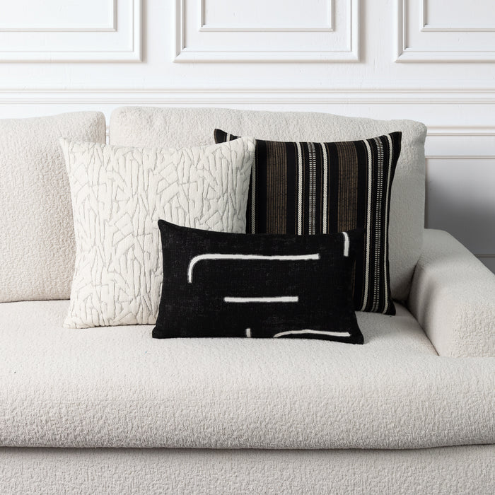 Three decorative throw pillows arranged on a textured off-white sofa against a white paneled wall. The front pillow is black with white abstract lines, the center pillow is cream with a raised abstract pattern, and the back pillow has vertical black, white, and brown stripes.