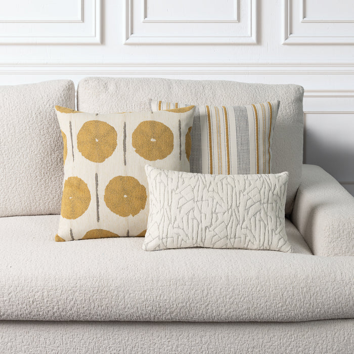 Three decorative throw pillows arranged on a light beige boucle sofa against a white paneled wall. The pillows include a cream pillow with mustard yellow circular motifs, a striped pillow in beige, gray, and yellow, and a cream textured lumbar pillow.