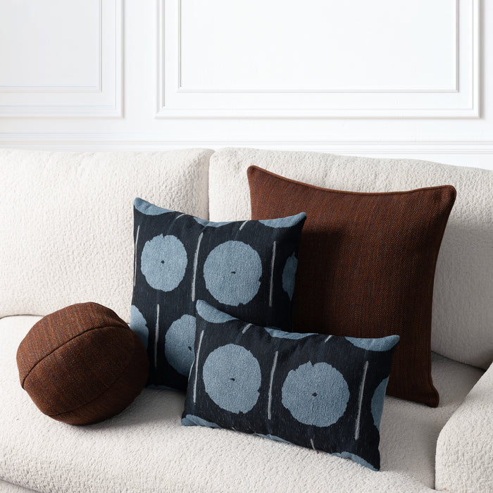 Three decorative throw pillows arranged on a cream boucle sofa against a white paneled wall. The set includes a round brown pillow, a square dark brown pillow, and two black pillows with a blue-gray circular ikat pattern.
