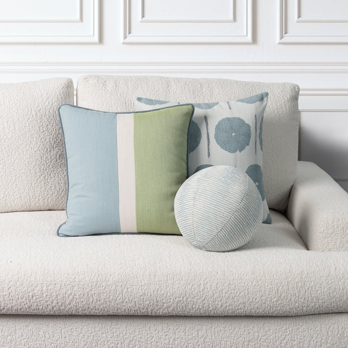 Three decorative throw pillows arranged on a textured cream boucle sofa against a white paneled wall. The front-left pillow is square with light blue, sage green, and off-white vertical stripes.