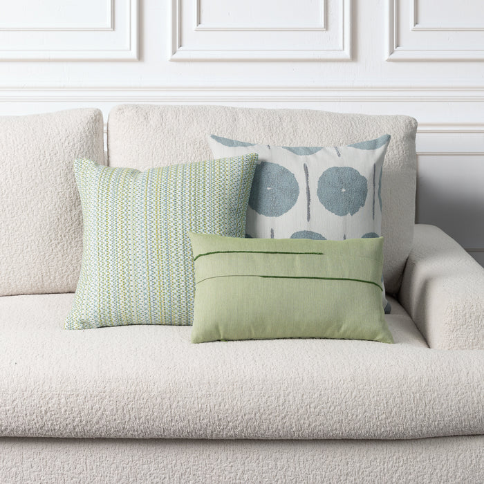 Three decorative throw pillows styled on a textured cream bouclé sofa against a white paneled wall. The pillows include a sage green lumbar pillow with horizontal stitching, a striped mint green and white pillow, and a white pillow with blue-gray circular patterns.