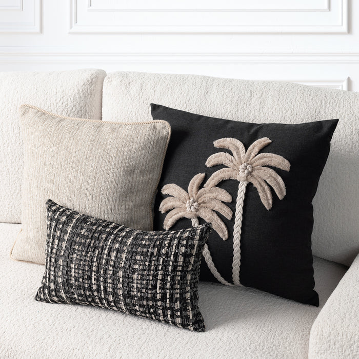 Three decorative throw pillows arranged on a cream textured sofa: a large black pillow with two beige palm tree motifs, a plain beige pillow, and a smaller black-and-beige woven lumbar pillow.